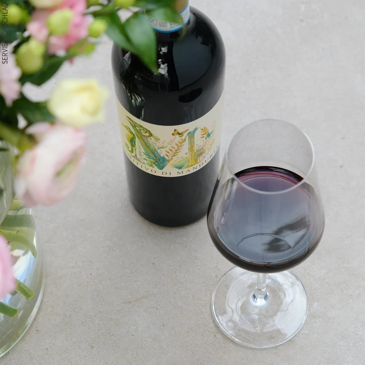 Der Primitivo di Manduria vereint süditalienische Kraft mit eleganter Finesse. Blumen, eine Flasche Primitivo di Manduria und ein Glas Rotwein